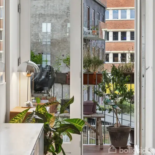 En balkon med planter i potter og grønne buske. Sollys strømmer ind gennem glasdøre, som fører til en indendørs stue med grønne planter og en bordlampe. Udsigt til bybygninger.