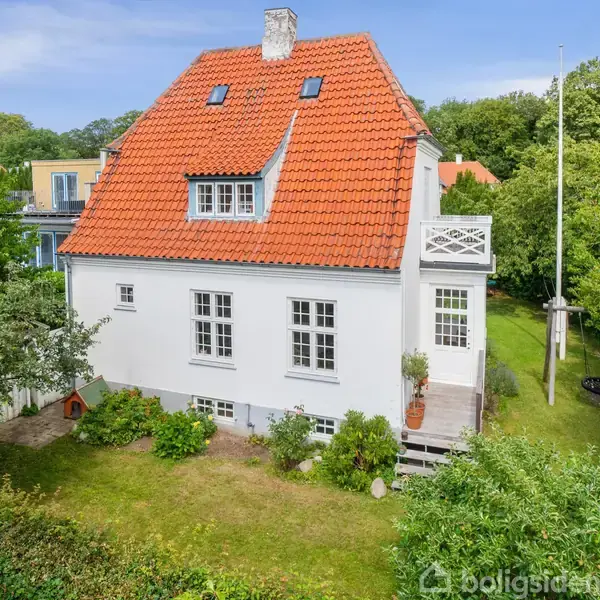 Hvidt hus med rødt tegltag står i en grøn have med buske og træer. En lille balkon og flagstang er ved siden af huset.
