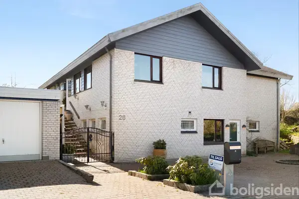 Hvid murstensvilla med mørke vinduer står stille i et solrigt nabolag. Skilt foran huset viser teksten "Til salg." Nummer 28 er synligt på muren.