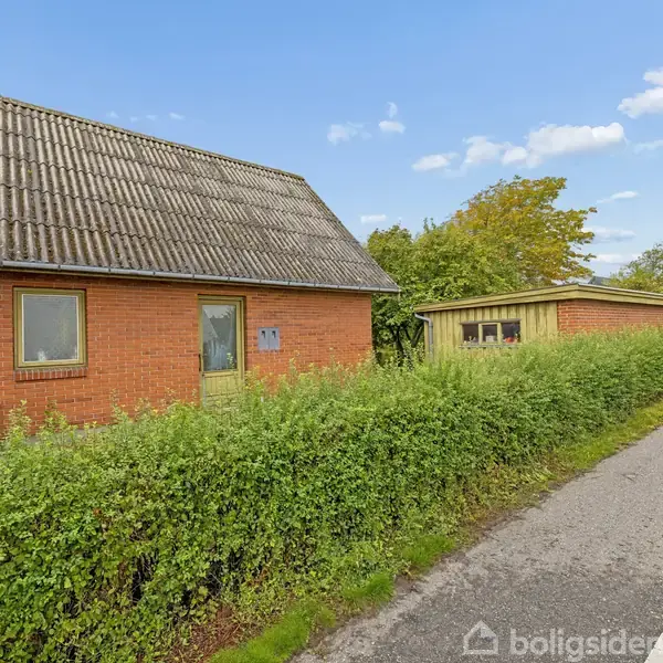 En rød murstensbygning med skråt tag står stille ved en landevej. En grøn hæk omgiver huset, og vejskilte ses i baggrunden under en blå himmel med få skyer.