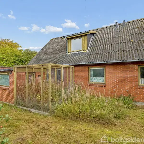 Murstenshus med skråt tag har et tilstødende drivhus omgivet af høj vegetation. Det ligger i en have med græs og buske under en blå himmel med skyer.