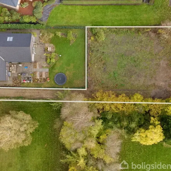 En rektangulær grund omgivet af vegetation, hvor et hus med en terrasse og en rund trampolin er placeret til venstre. Den højre sektion er en åben græsmark.