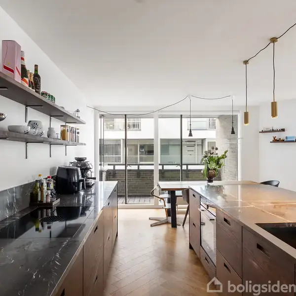 Køkken med mørke skabe og marmor bordplader med en vask og guldhane. Hylder over bordpladen bærer glas og redskaber. Store vinduer giver udsigt til en balkon.