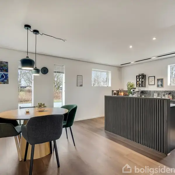 Rundt spisebord med fire stole, placeret i en moderne spisestue. Til venstre ses tre kunstværker på væggen, mens der til højre står et stilfuldt køkken med mosaikfliser og vinduer.