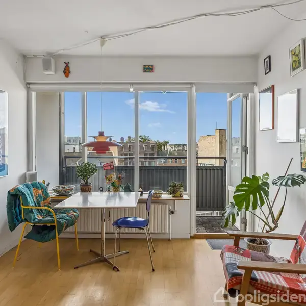 Stue med lyse træmøbler, hvor to stole og et bord er placeret ved en stor glasdør, der åbner ud til en balkon. Indretningen er farverig med planter og kunstværker.