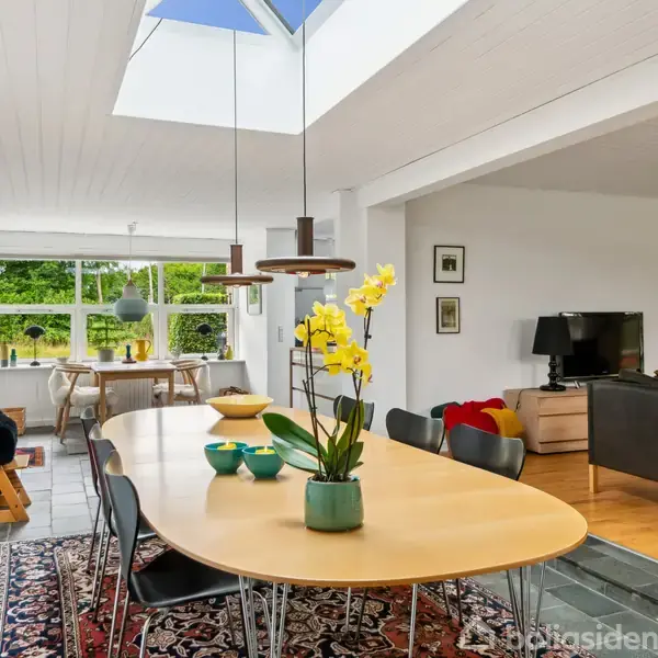 Et moderne spisebord med gule blomster i en vase står i centrum af et lyst rum. På væggen hænger billeder, og i baggrunden ses sofa og spiseområde foran vinduer.