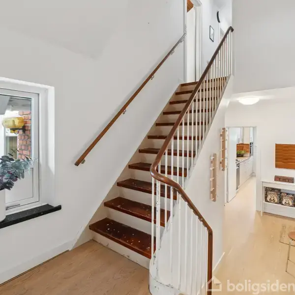 Trappe med trætrin og hvidt gelænder fører opad. Ved siden af er et vindue med potteplanter. Gangen er lys med åbning til et køkkenområde.