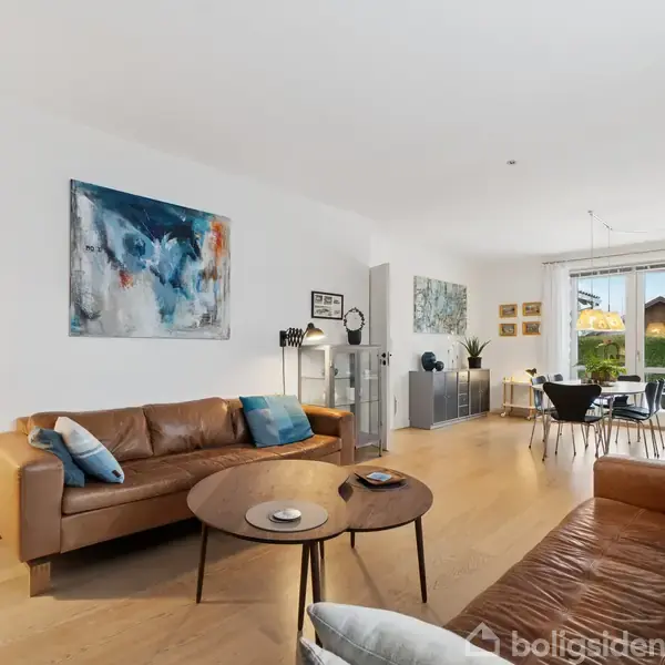 To lædersofaer står i en lys stue med trægulv og kunst på væggene. Et sofabord er centralt placeret, og et spisebord ses i baggrunden nær vinduerne.