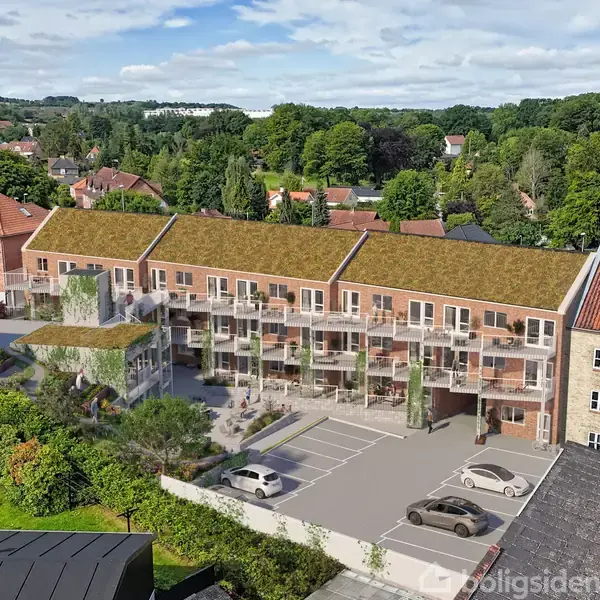 Rækkehuskompleks med grønne tage omgivet af parkeringspladser og frodig vegetation, set fra en fugleperspektiv. Baggrunden viser et skovklædt landskab og flere bygninger.