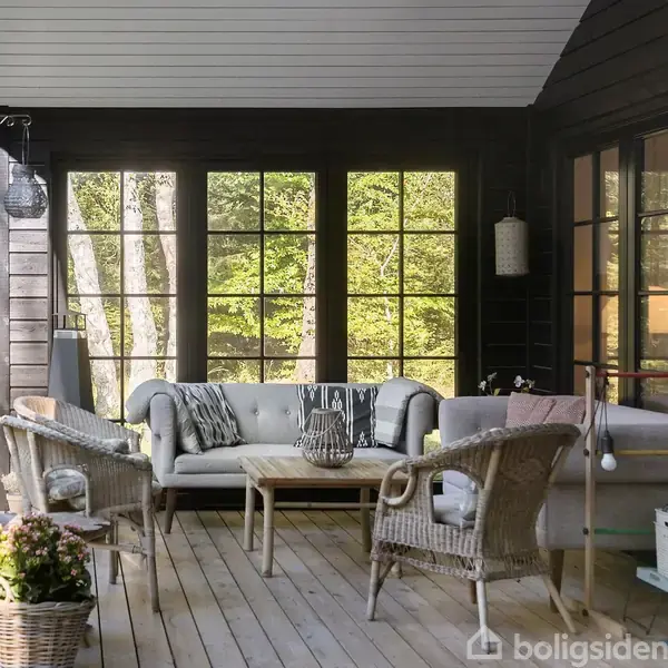 En overdækket terrasse med polstrede møbler og et sofabord, hvor hoveddøren står åben. Omgivet af glasvægge med udsigt til en grøn have og skov.
