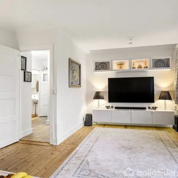 TV ophængt på væg, placeret over en hvid skænk med lamper, i en stue med lyst trægulv og tæppe. Væghylder med dekoration over. Vindue med lange gardiner til højre.