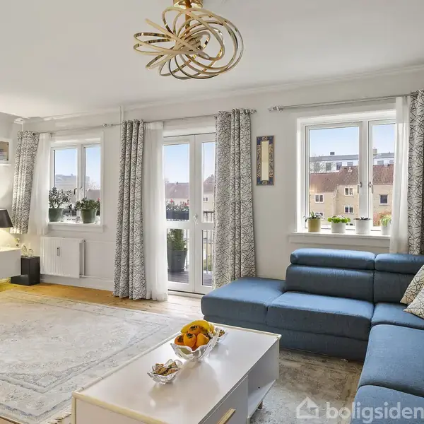 Stor, lys stue med blå hjørnesofa placeret foran et sofabord med frugtskål. Vindueselementer med gardiner lukker lys ind, mens en fladskærm hænger på væggen. Elegant loftslampe fuldender rummet.