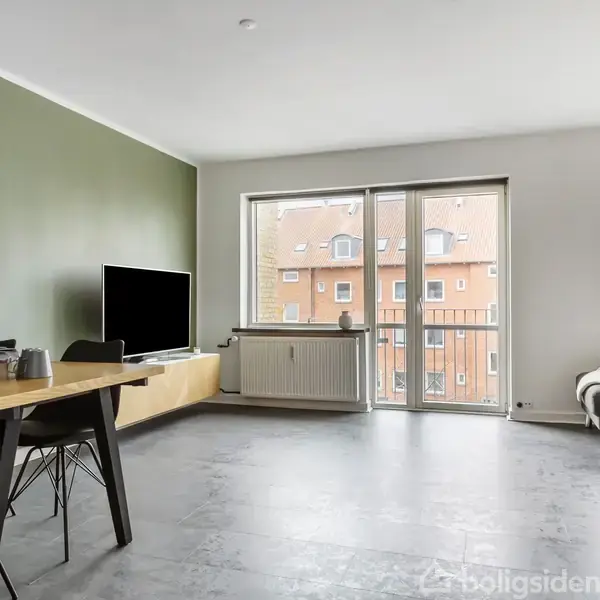 Stue med tv på skænk ved grøn væg. Langt bord med sorte stole til venstre. Store vinduer giver udsigt til murstensbygning. Lys grå sofa står ved siden af.