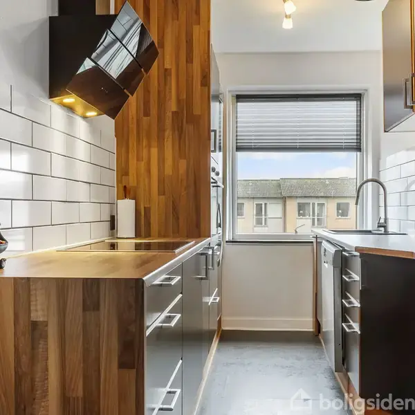 Et moderne køkken med mørke skabe og træbordplader. Køkkenet har en indbygget komfur og hvid vægflisebaggrund. Et stort vindue giver udsyn til bygninger udenfor.