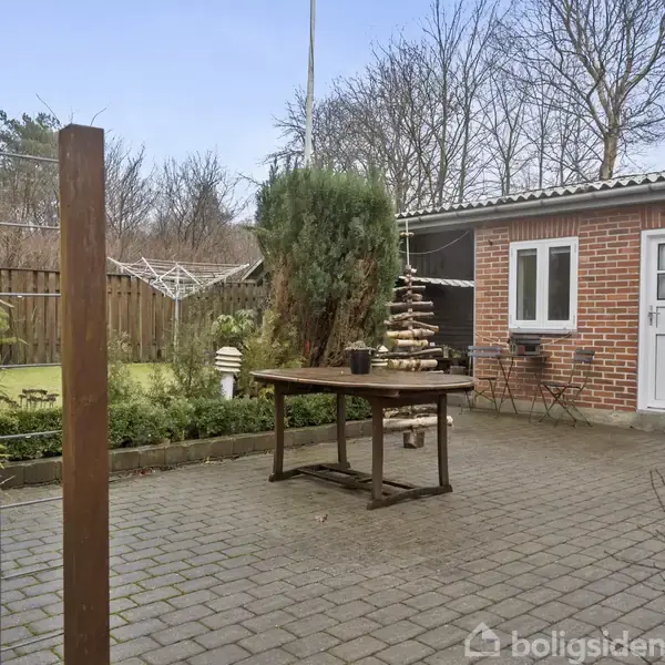 Et træbord står på en brolagt terrasse foran en murstensbygning med en hvid dør. I baggrunden ses en grøn have med høje træer og en træhegn.