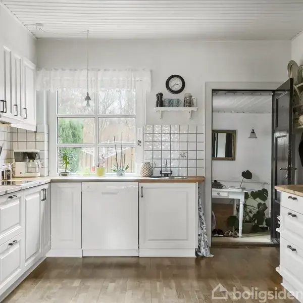 Køkken med hvide skabe og træbordplader, står stille under et ur. Sollys skinner ind gennem vinduerne, mens dør fører til rum med planter og spejl.