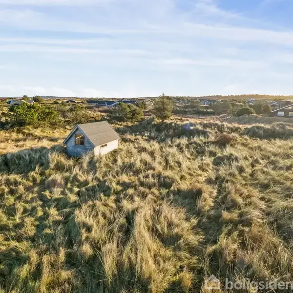 Et lille hus står alene i en stor, åben mark med højt græs. Omgivet af spredte træer og andre små bygninger i det fjerne, under en klar blå himmel.