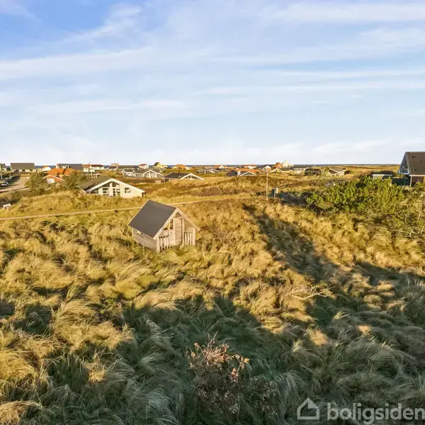 En lille hytte står stille omgivet af høje græsmarker. I baggrunden ses flere huse og en klar blå himmel, der antyder et fredeligt landskab.