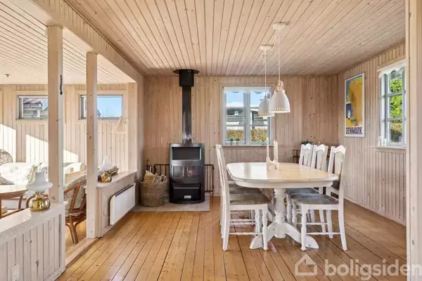 Et træspisebord med seks stole står centralt i en stue med brændeovn og lyse trævægge. Der hænger lamper over bordet og en plakat med teksten "DENMARK" på væggen.