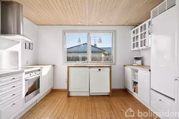 Et lyst køkken med hvide skabe og træloft. Der er en ovn til venstre og en vindue i midten mod en fliseterrasse. Trægulve fuldender det moderne udseende.