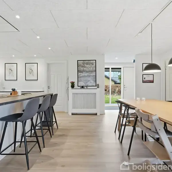 Et moderne køkken-alrum med høje stole ved en mørk køkkenø og en lang spisebord med stole. Lyse trægulve og hvide vægge med kunst hængende.