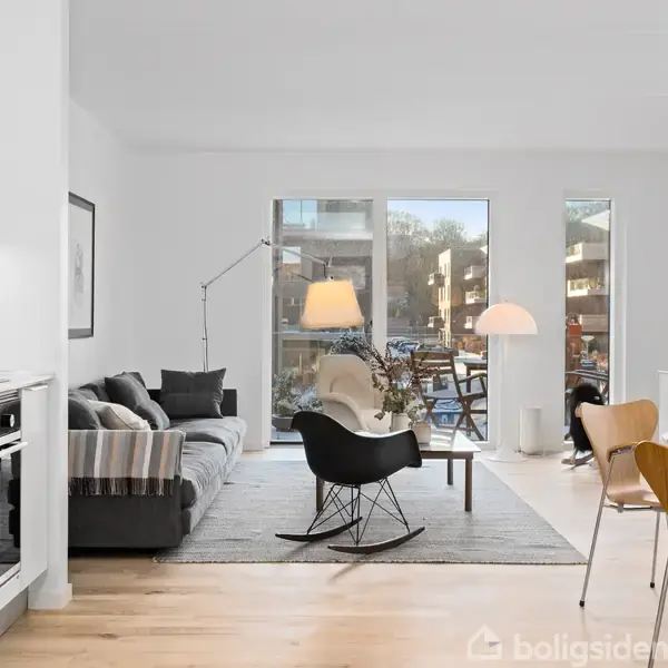 En moderne stue med en grå sofa, gyngestol og spisebord, oplyst af naturligt lys fra store vinduer, der vender ud mod en terrasse. Køkkenområdet er til venstre.