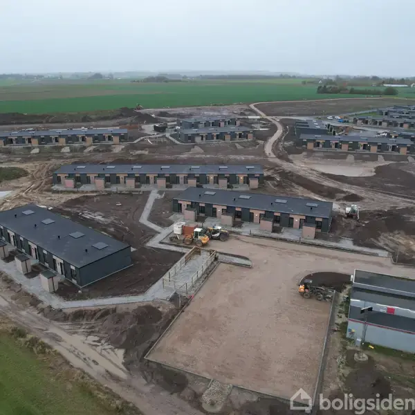 Flere rækker af nybyggede huse strækker sig gennem et anlægsområde. Byggepladsen har spredte maskiner og containere. Omkringliggende marker og veje strækker sig i horisonten.