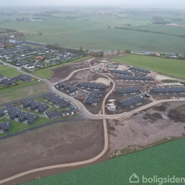 Byområde under udvikling med nye boligkomplekser omgivet af åben landbrugsjord og eksisterende huse. Anlægsarbejde kan ses med veje og fundamenter i forskellige byggestadier.