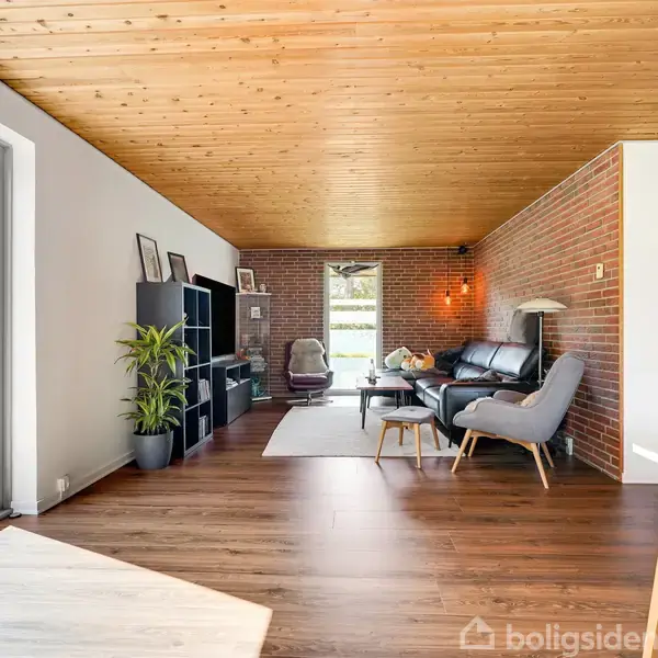 Stue med sort lædersofa og grå stol står på et parketgulv med tæppe. Rød murstensvæg i baggrunden, moderne reol, store vinduer og træloft. Udgang til træterrasse på venstre side.