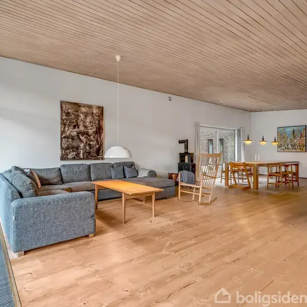 Stue med et langt spisebord og stole, står i et rummeligt rum med trægulve og loft, store vinduer og en sofa i baggrunden. Store glasdøre giver udsigt til haven.