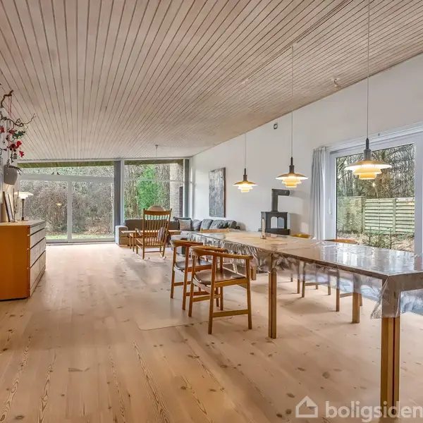 Stort rum med spisebord og stole placeret på trægulv. Store vinduer giver udsigt til en have med høje træer og buske. Moderne lamper hænger fra loftet.