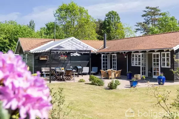 Et hyggeligt sommerhus med rødt tegltag står i en have med græsplæne og blomster. Havemøbler er arrangeret på terrassen. Skoven omkranser området i baggrunden.