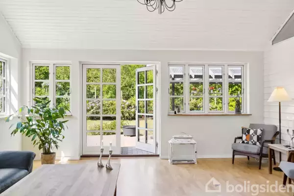 Sorte lædersofaer står i en lys stue med trægulv. Et træbord står i midten. Store vinduer og glasdøre giver udsigt til en grøn have. En lysekrone hænger fra loftet.