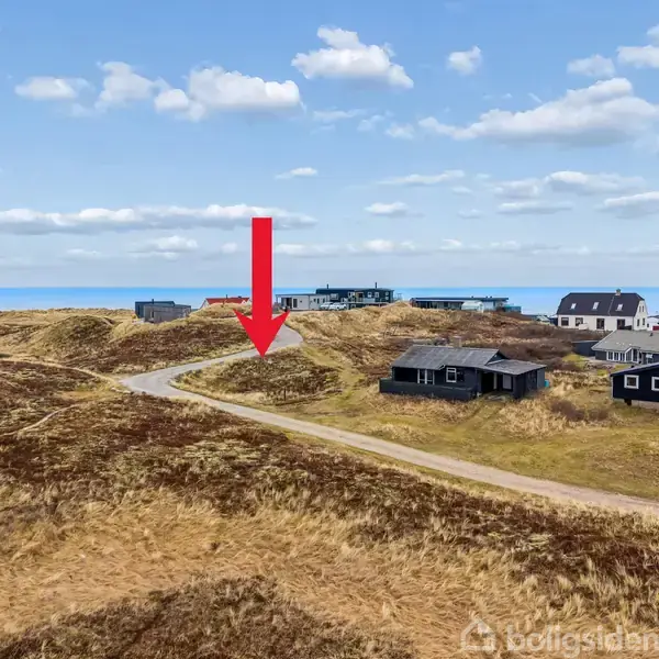 Røde pil peger mod vej gennem græsbeklædte klitter. Sorte og hvide huse ligger spredt omkring. Hav og blå himmel med skyer i baggrunden.