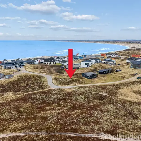 En rød pil peger mod et hus i et kystnært boligområde. Omkringliggende huse ligger spredt på en bakke med havet i baggrunden. Skydække er let med spredte skyer.
