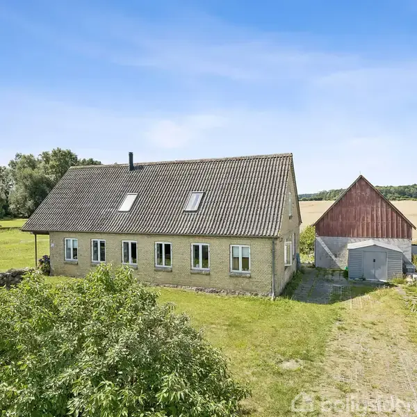 Et gult murstenshus med kvist står på en stor græsplæne ved siden af en rød lade. Flagstang uden flag i forgrunden; marker og skov i baggrunden.