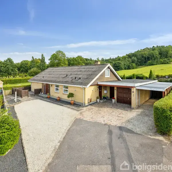 Et gult murstenshus med to garager ved siden. Huset står stille i en landskabelig, landlig omgivelser med frodig grøn vegetation og bakker i baggrunden.