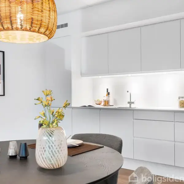 Et rundt spisebord i forgrunden med en vase med blomster og service. En simpel, moderne køkkenindretning i baggrunden med hvide skabe og en væglampe. En flettet lampe hænger over bordet.