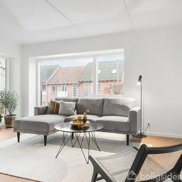 Stue med grå sofa ved stort vindue, sofabord med dekorationer, og en stol i forgrunden. Lys lampe ved sofaen og planter nær altandør. Lys og moderne indretning.