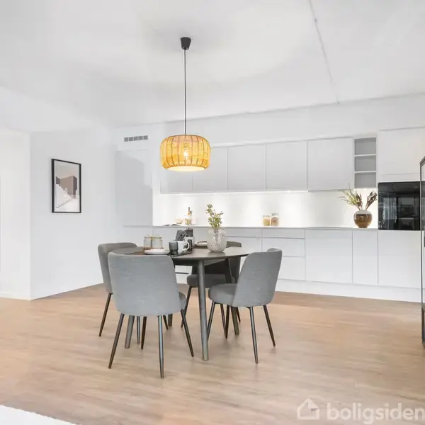 Spisebord med fire stole står i midten af et moderne, hvidt køkken med skabsvæg og hvidevarer. En pendellampe hænger over bordet, og en reol står til højre.