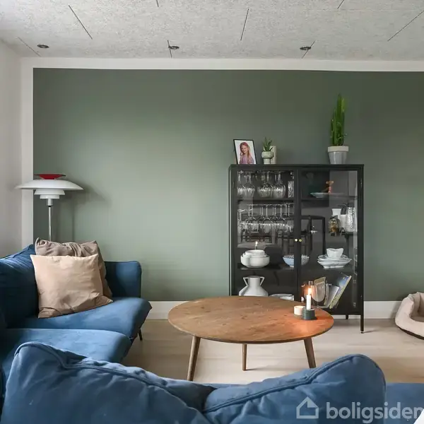 En blå sofa står i en moderne stue med grøn accentvæg. Et træbord er midtpunkt, omgivet af en glasvitrine med porcelæn. En lampe og planter tilføjer hygge.