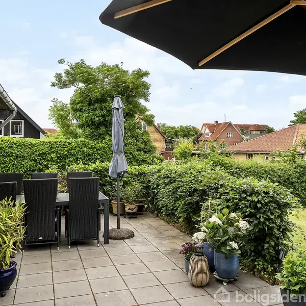 Terrasse med sorte havemøbler under en lukket parasol; omgivet af tætte grønne buske og planter. I baggrunden ses hustage og en blå himmel.