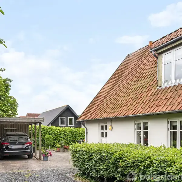 Et hvidt hus med rødt tegltag står ved siden af en carport med en parkeret bil. Grønt løv omgiver indkørslen, og himlen er klar og blå.