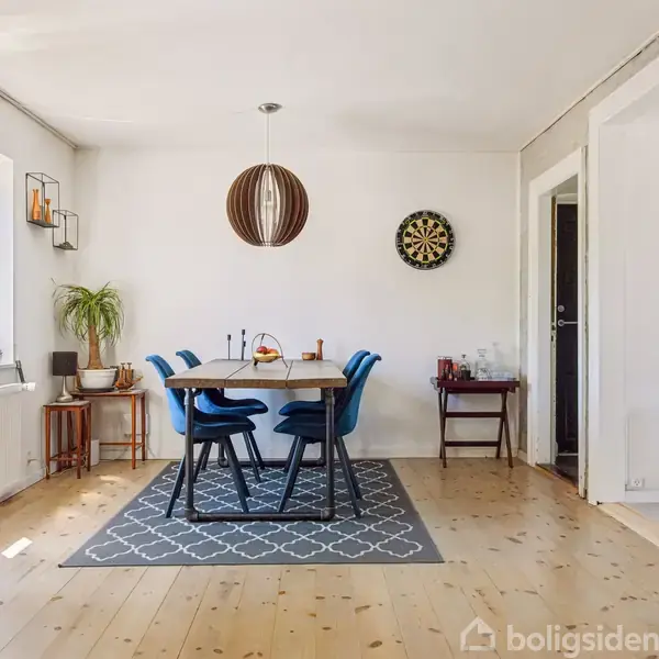 Et spisebord med blå stole står midt i et lyst rum. En lampe hænger over bordet. En dartskive hænger på væggen. Tekst: "VERDENS BEDSTE FAR TALJENS".