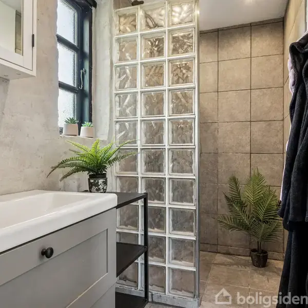 Håndvask står under spejlskab i badeværelse med beige vægge. Flere planter dekorerer rummet. Bruseniche med glasbyggesten og fliser. Mørke håndklæder hænger på højre væg.