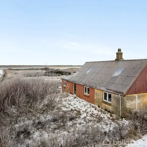 Et gammelt hus står tomt tæt ved et kystområde omgivet af buske og græs. Himlen er klar, og der er en vandflade i baggrunden.