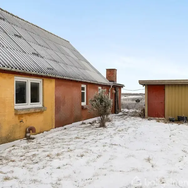 Et ældre hus med skråt tag står i et snedækket, åbent område. Til siden er en lille skurbygning. Forgrunden er dækket af let sne.