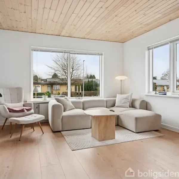 En lys stue med en beige hjørnesofa står ved vinduer, der lukker dagslys ind. En lænestol er til venstre, omgivet af træmøbler og planter.