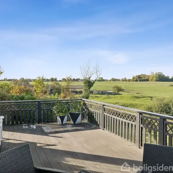 En terrasse med sorte og hvide havemøbler strækker sig ud i en landlig omgivelser med udsigt over en grøn mark under en klar blå himmel.