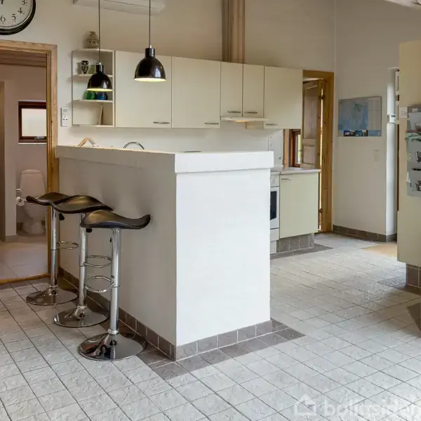 Køkken med to barstole ved en hvid bardisk. Rummet har hængende lamper, køkkenskabe og synligt ur. En åben dør fører til et badeværelse. Lys fylder rummet.